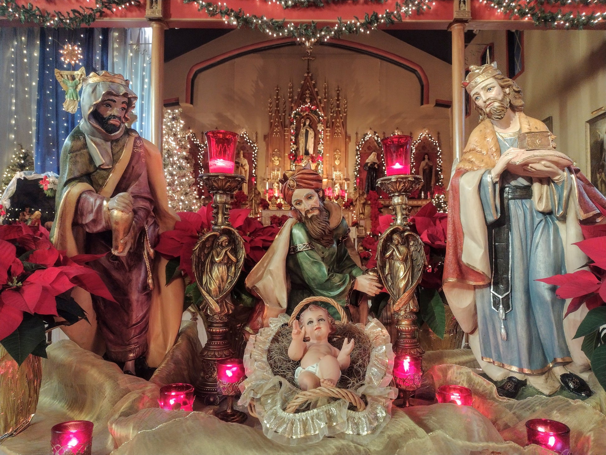 Nativity scene with three wise men and baby Jesus in a manger, surrounded by poinsettias and candles.